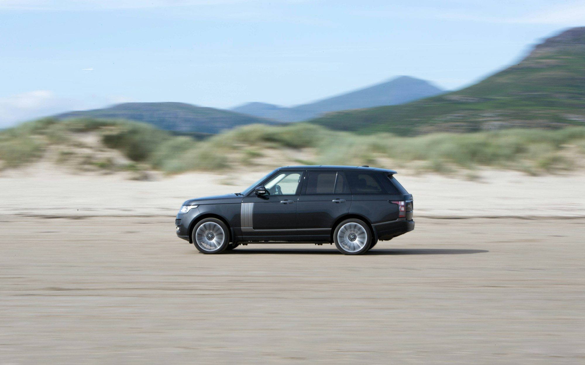 A black fourth-generation Range Rover (L405) driving across a sand beach with dunes and hills behind