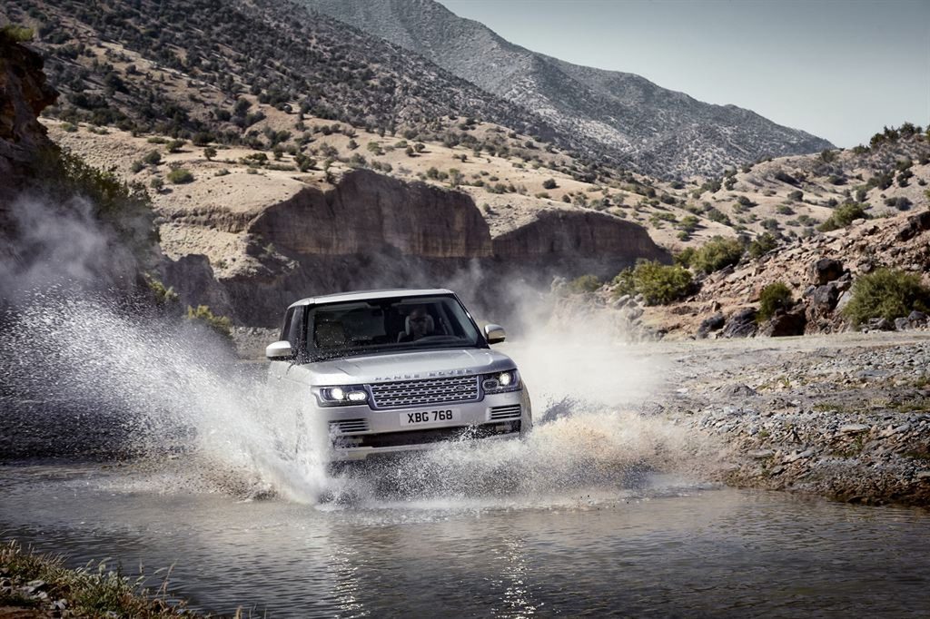 A silver pre-facelift L405 Range Rover splashing through a shallow river in a rocky mountain valley