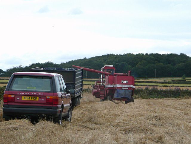 A red P38A Range Rover parked in a stubble field next to a grain trailer and combine harvester