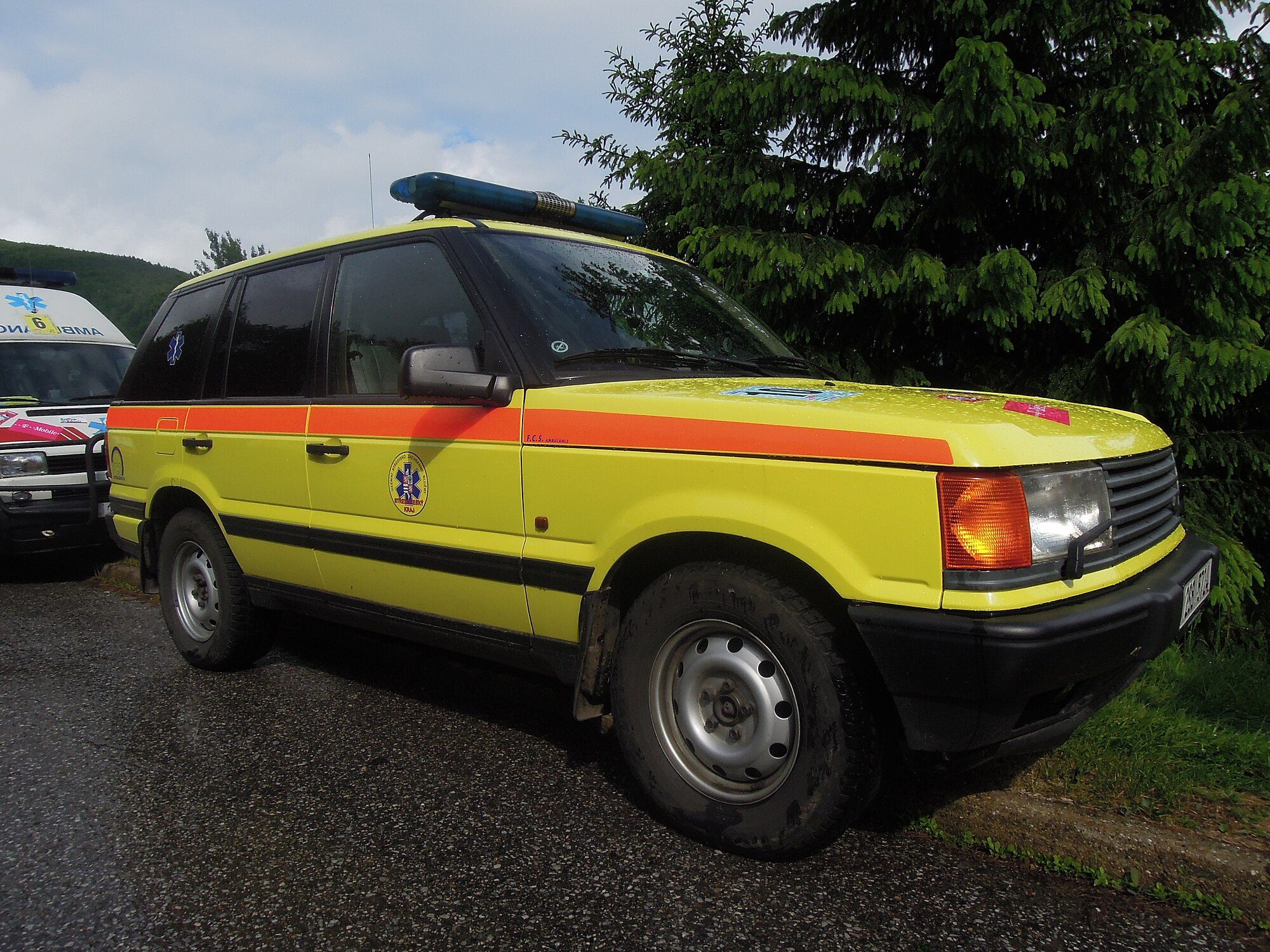 A yellow second-generation Range Rover (P38A) ambulance parked on a wet tarmac lane beside spruce trees at the Rejvíz rescue rally