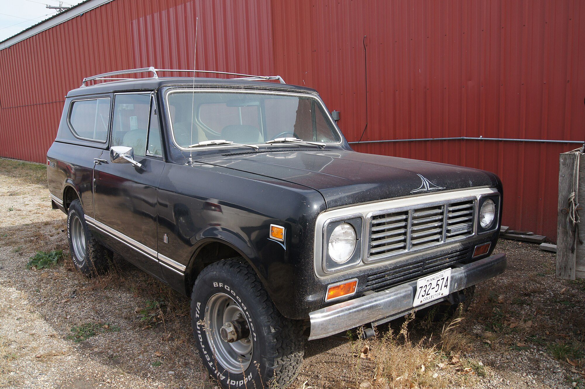 A black 1976 Scout II with roof rack and all-terrain tires parked on gravel beside a red farm building