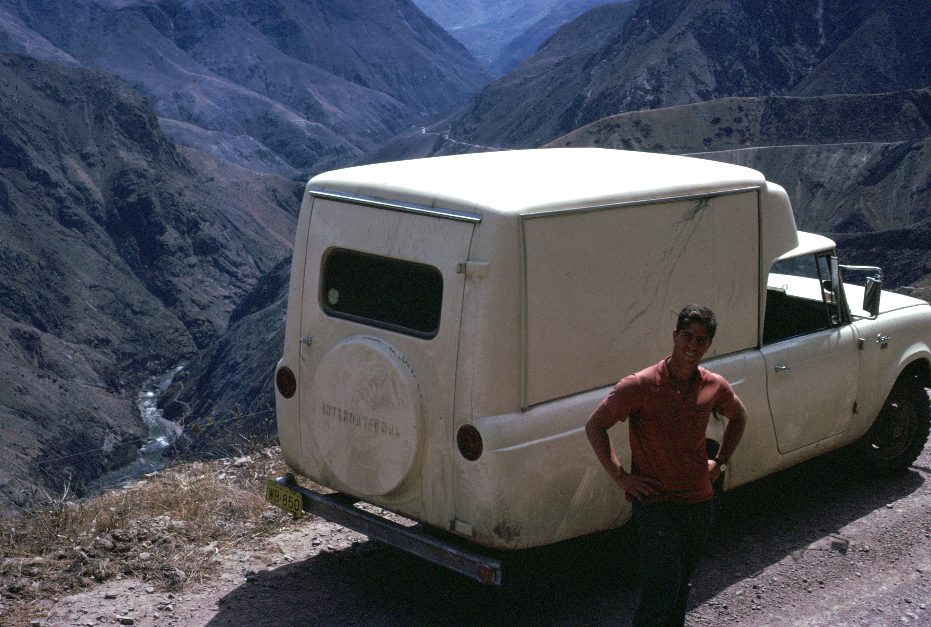 A white 1963–64 Scout 80 Camper parked on a dirt mountain road above a deep river canyon, with a young man standing beside it