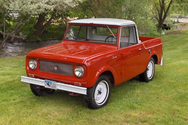 A restored red 1961 International Scout 80 pickup with a white top parked on grass beside a stream