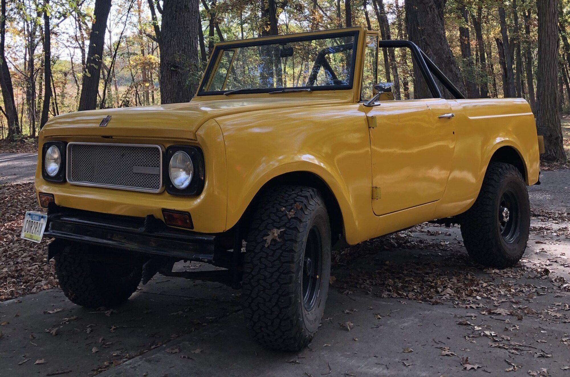 A yellow open-top International Scout 800-series (the uploader's file name says 800A) with a roll bar and all-terrain tires on a leaf-covered paved lane in autumn woods in Iowa