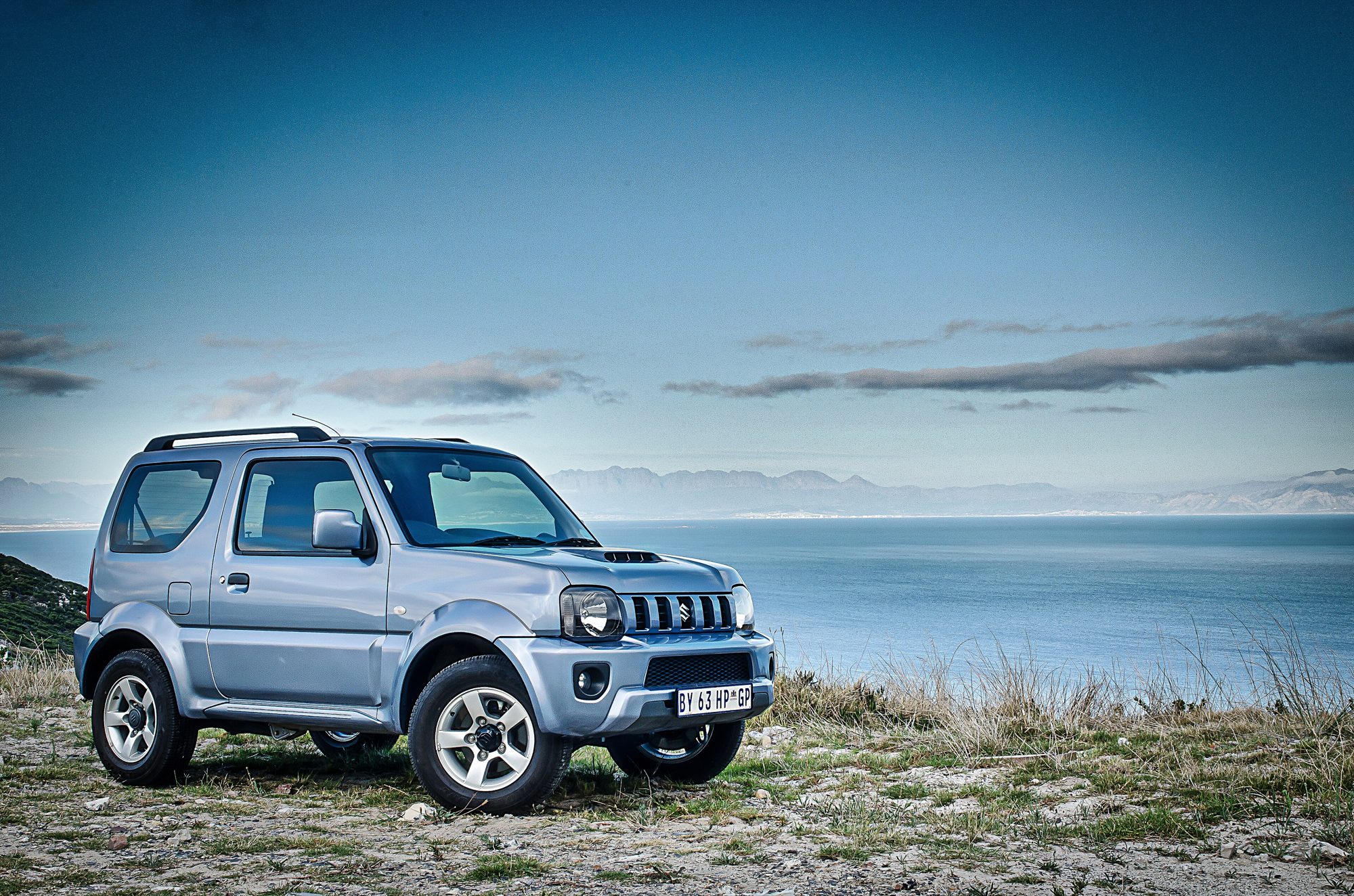 A silver late-model third-generation Suzuki Jimny with a hood scoop parked on a rocky clifftop above the sea