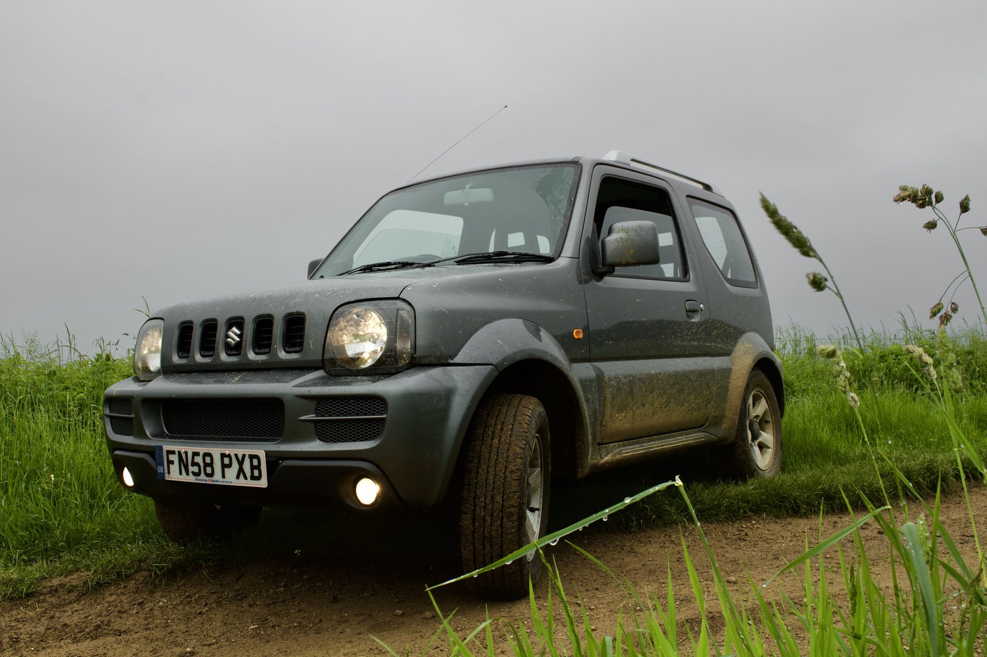 A muddy grey Suzuki Jimny 1.3 JLX+ on a dirt track between tall grass in Wiltshire under an overcast sky