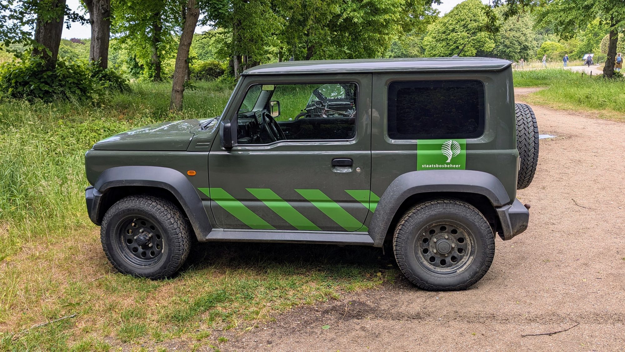 A dark green Staatsbosbeheer forestry Suzuki Jimny on steel wheels parked on a dirt path at the edge of a Dutch woodland