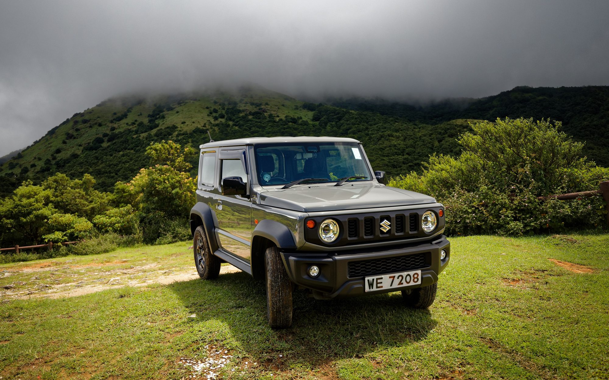 A grey fourth-generation Suzuki Jimny parked on a grassy hillside below misty green mountains