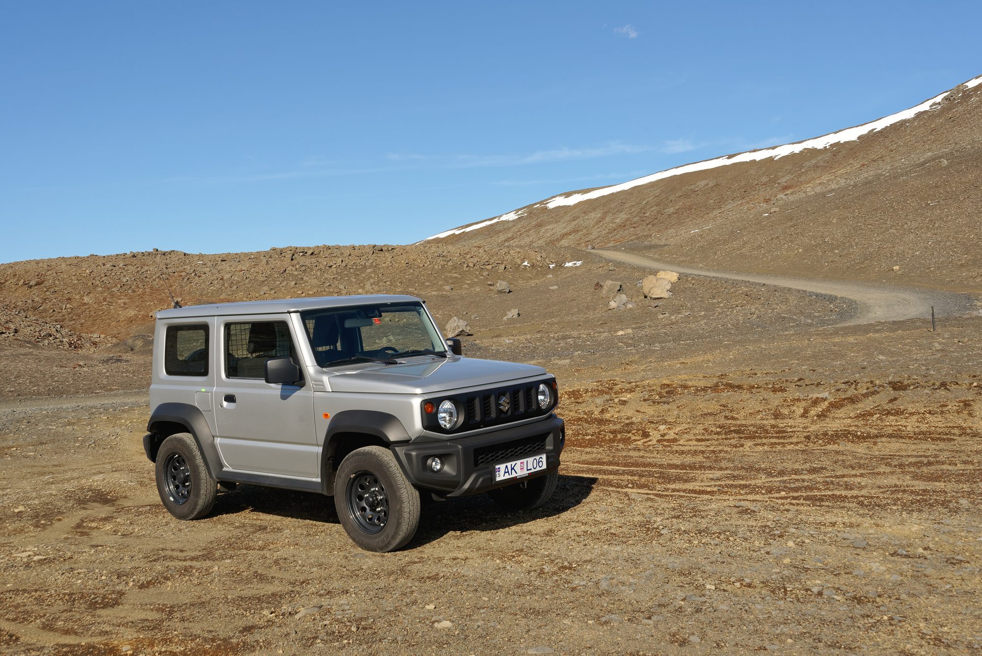 A silver fourth-generation Suzuki Jimny on a dirt track in the barren volcanic Fagradalsfjall mountains of Iceland, with snow on the ridge behind