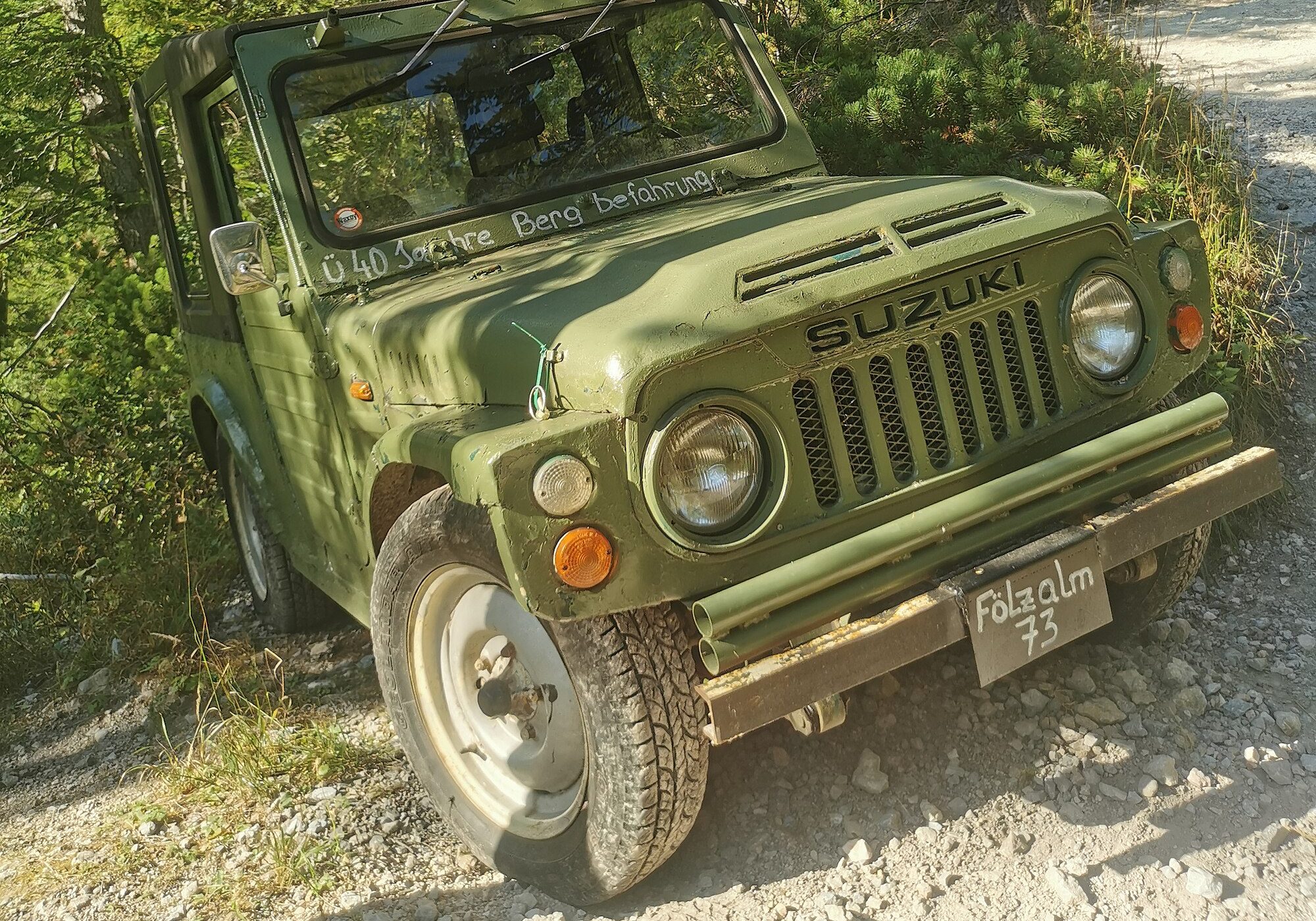 An olive-green 1973 Suzuki Jimny LJ20 on a gravel mountain track among larch trees, still used to supply an Austrian mountain hut