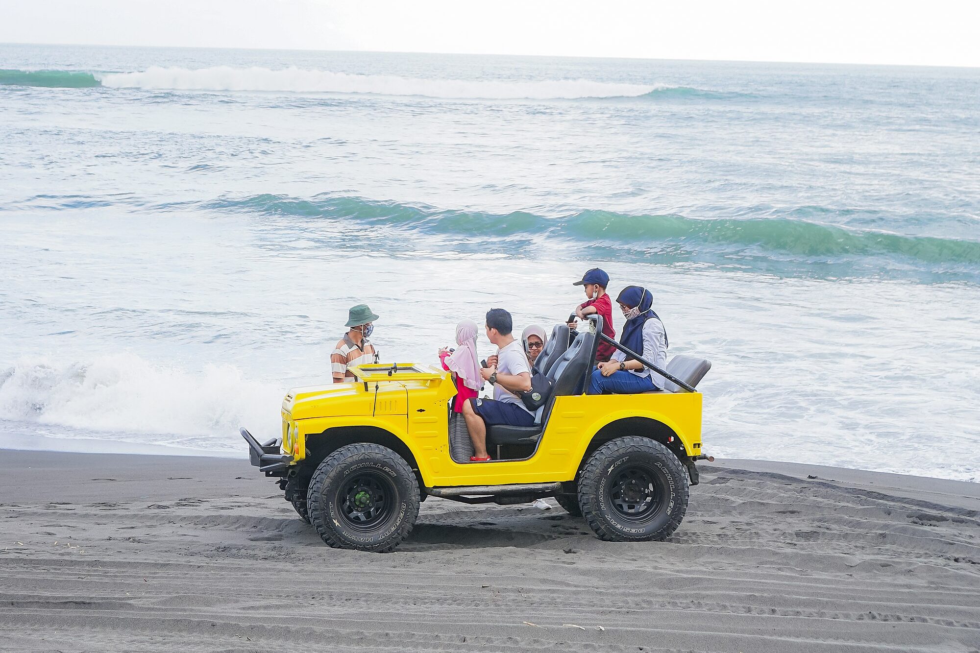 A yellow open Suzuki LJ80 carrying a family along a black-sand beach near Yogyakarta, Indonesia