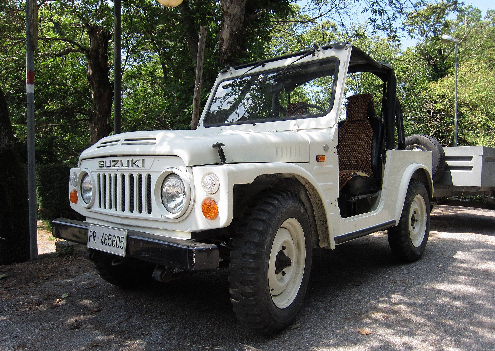 A white 1981 Suzuki LJ80 soft top parked on gravel under trees in rural Tuscany