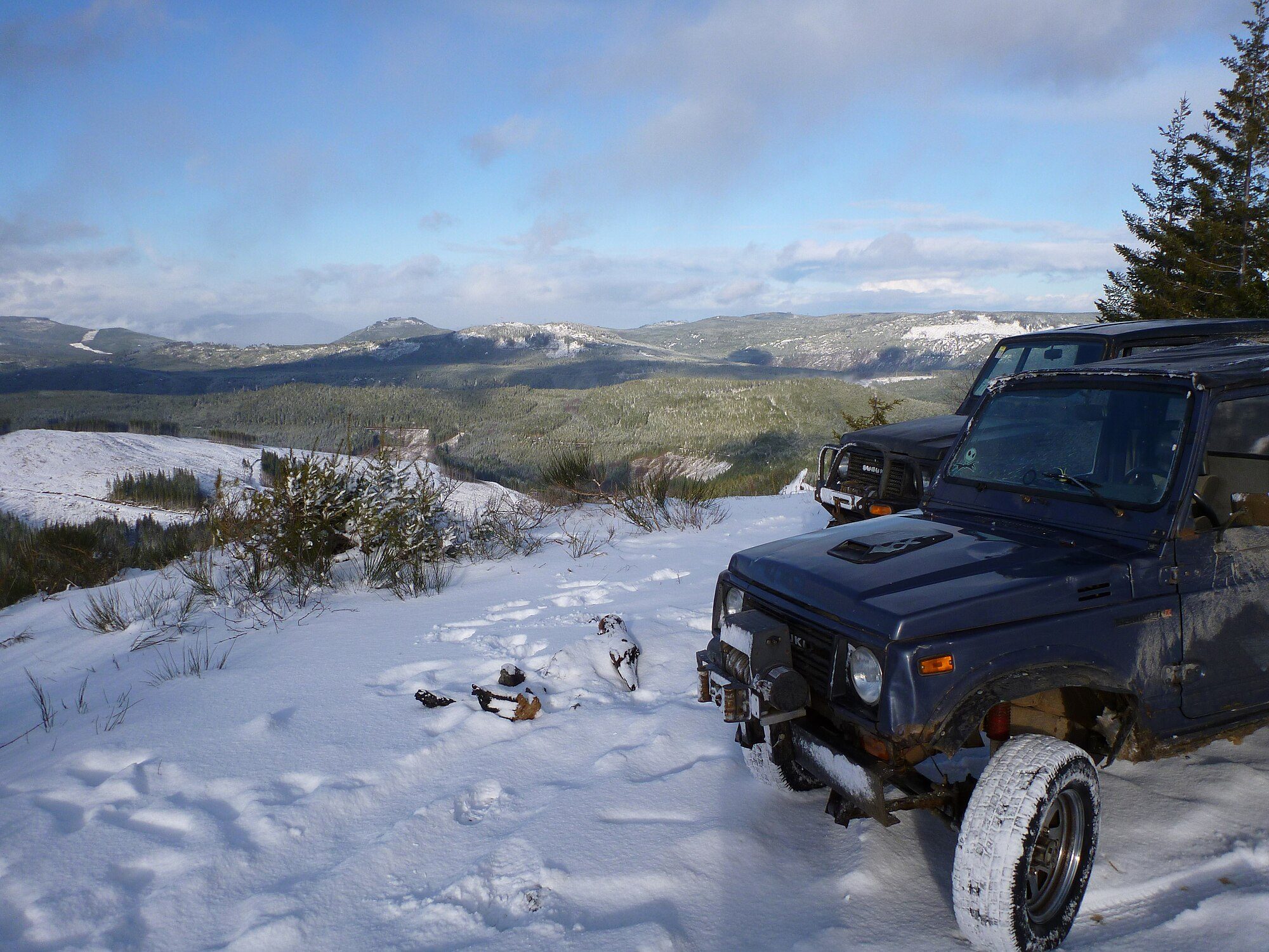 A dark blue Suzuki Samurai with a winch parked in fresh snow on a mountain ridge overlooking forested valleys