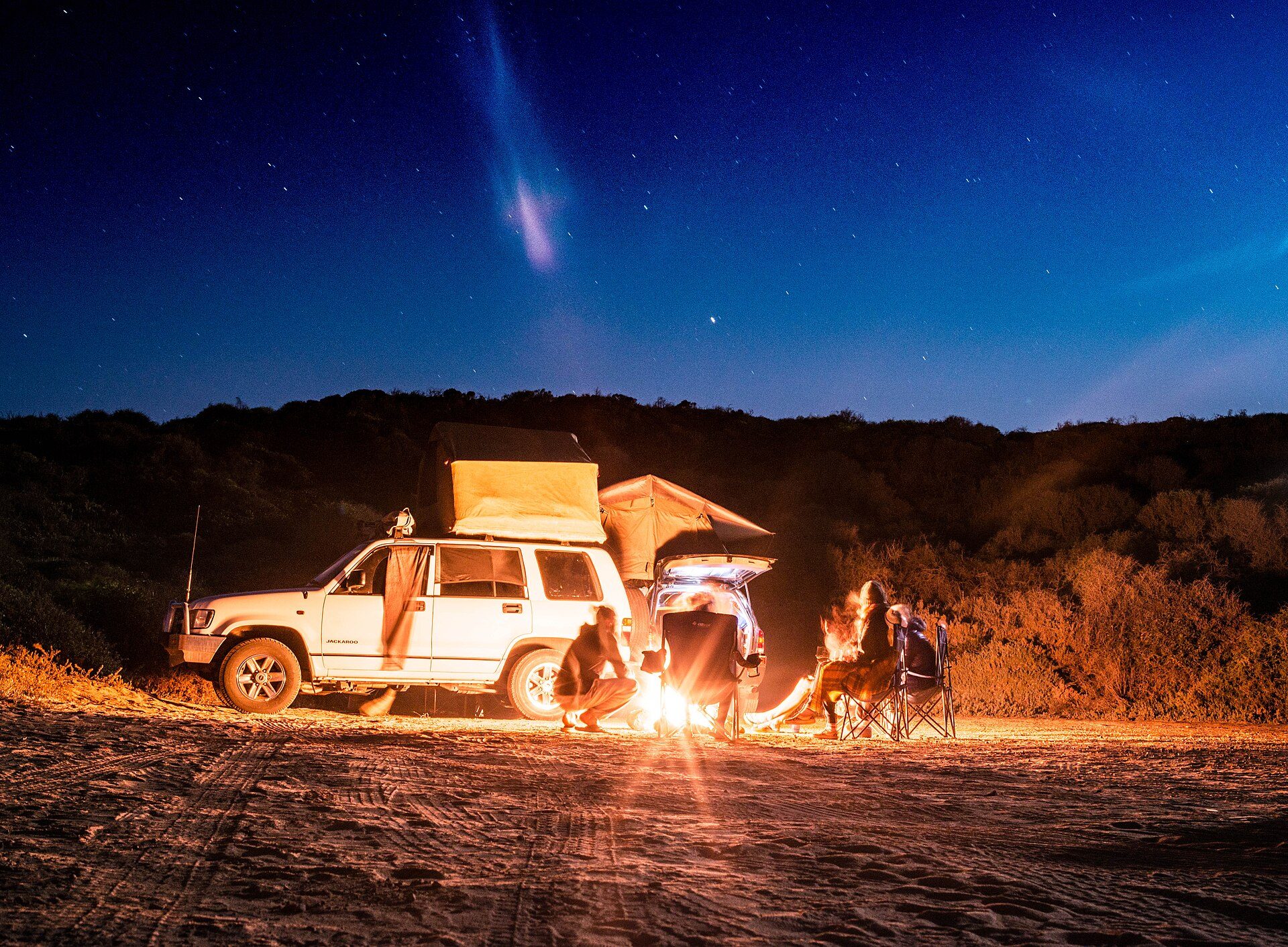 A white second-generation Holden Jackaroo with rooftop tents at a sandy beach camp at night, people around a campfire under a starry sky