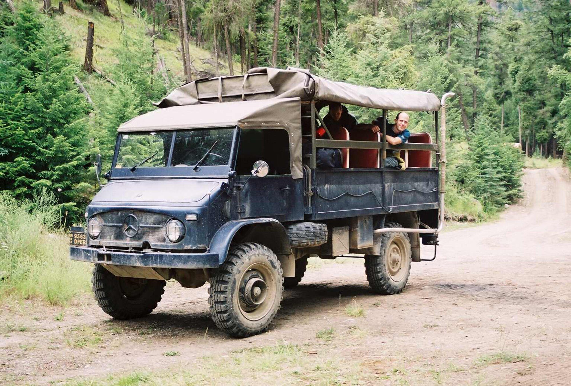 A dark blue Unimog 404.1 troop carrier with a canvas top and passengers in the back, on a forest dirt road