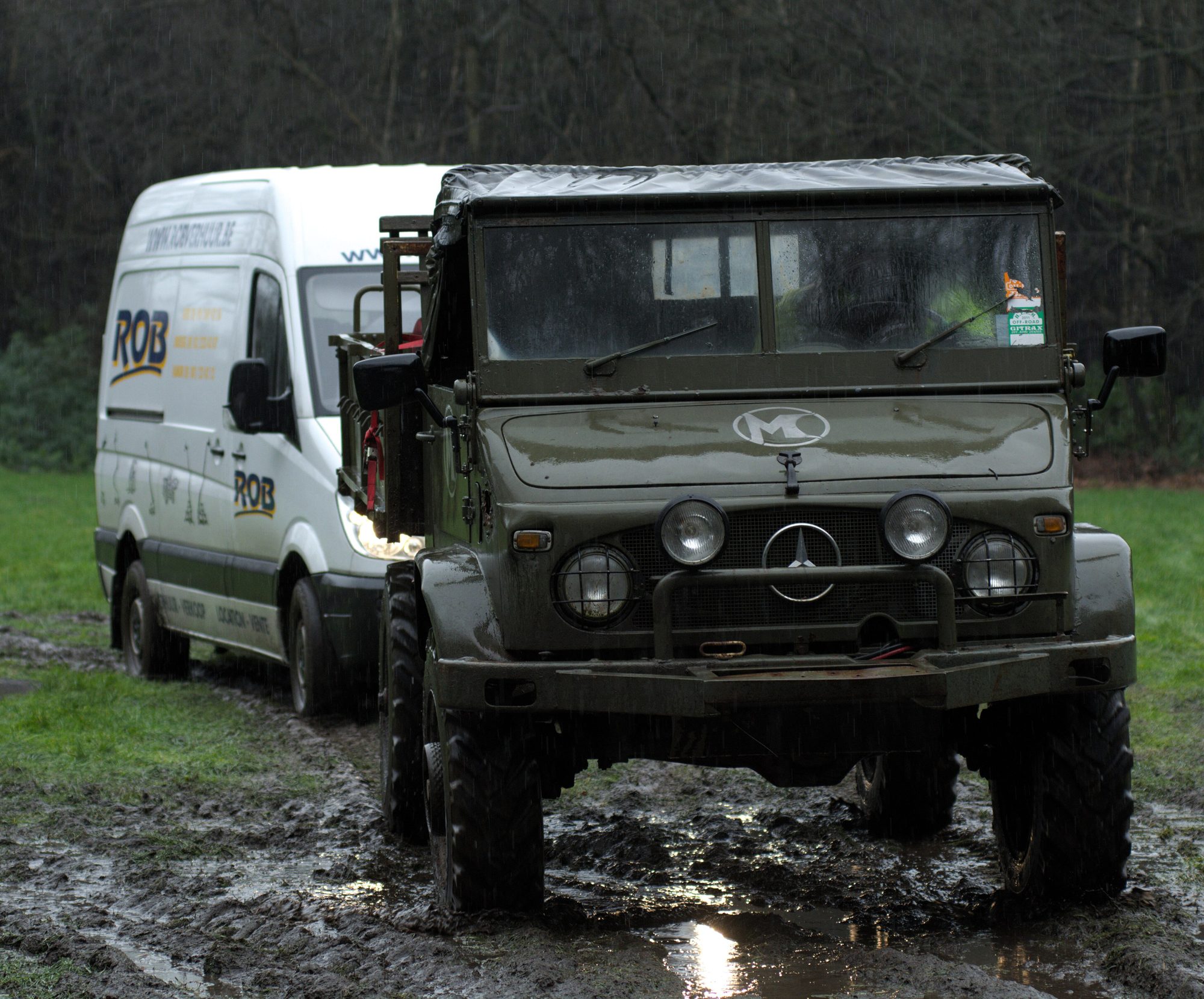 An olive-green Unimog 404.1 with a canvas top standing in a muddy field in the rain, a van behind it