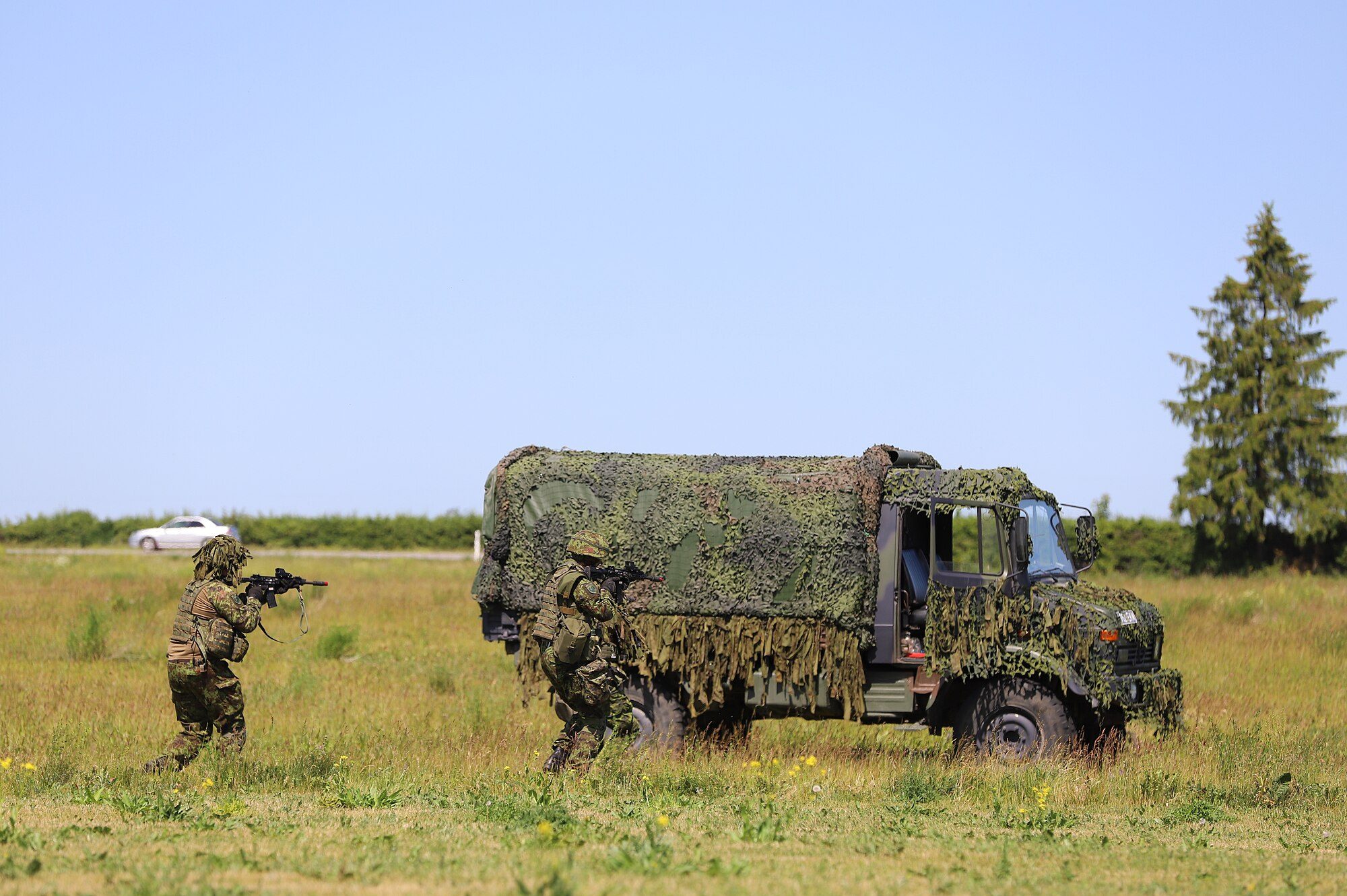 Two soldiers with rifles advancing past a camouflage-netted Unimog U1300L parked in a meadow