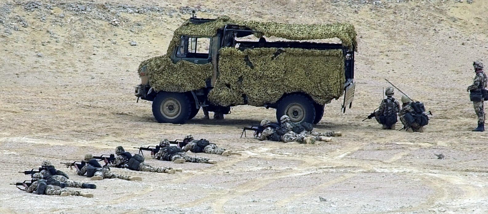 A German Army Unimog U1300L draped in desert camouflage netting on sandy, rocky ground while soldiers lie prone in front of it