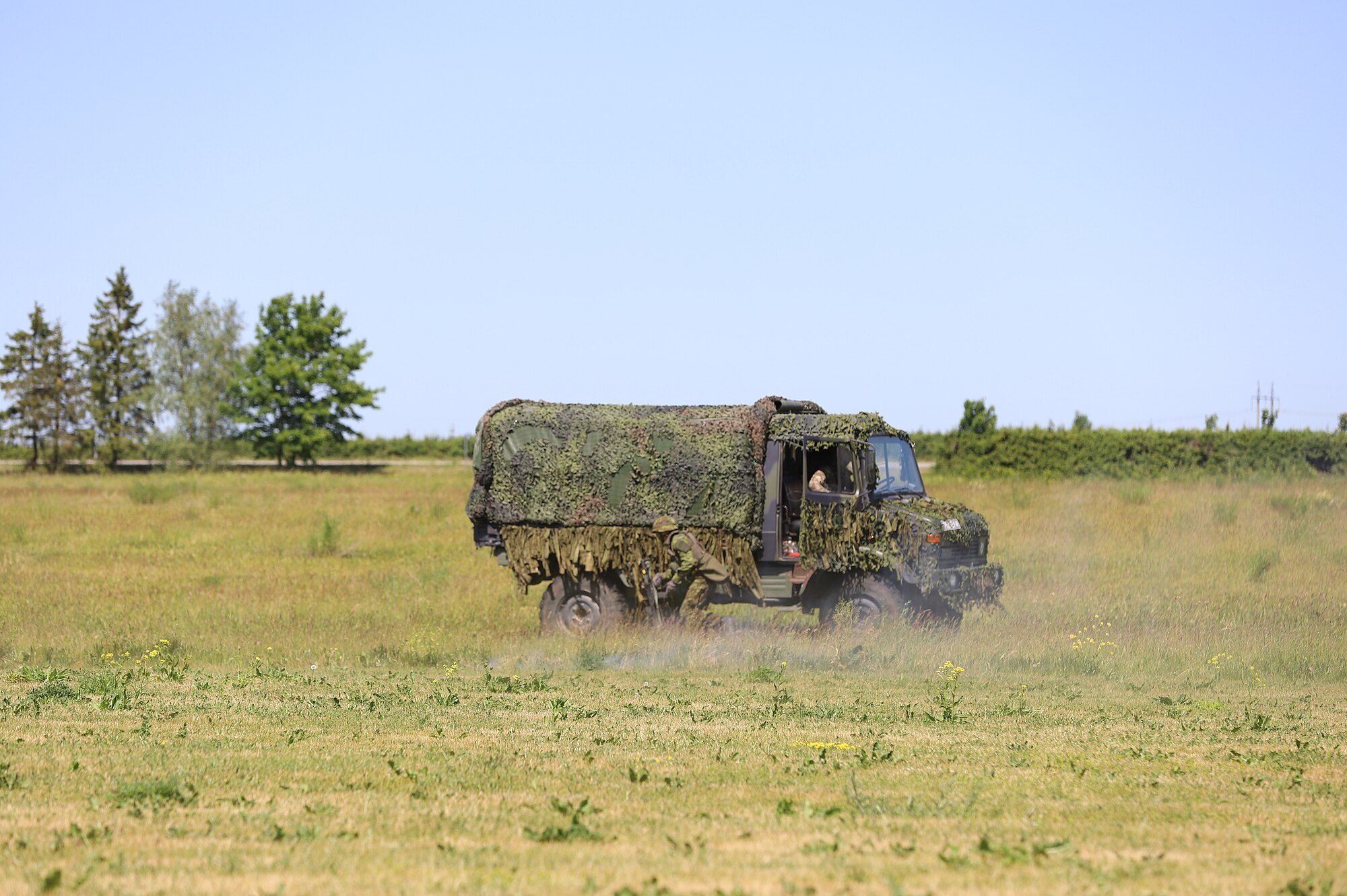 A camouflage-netted Unimog U1300L cargo truck driving across a grassy field, kicking up dust, with a soldier crouched beside it