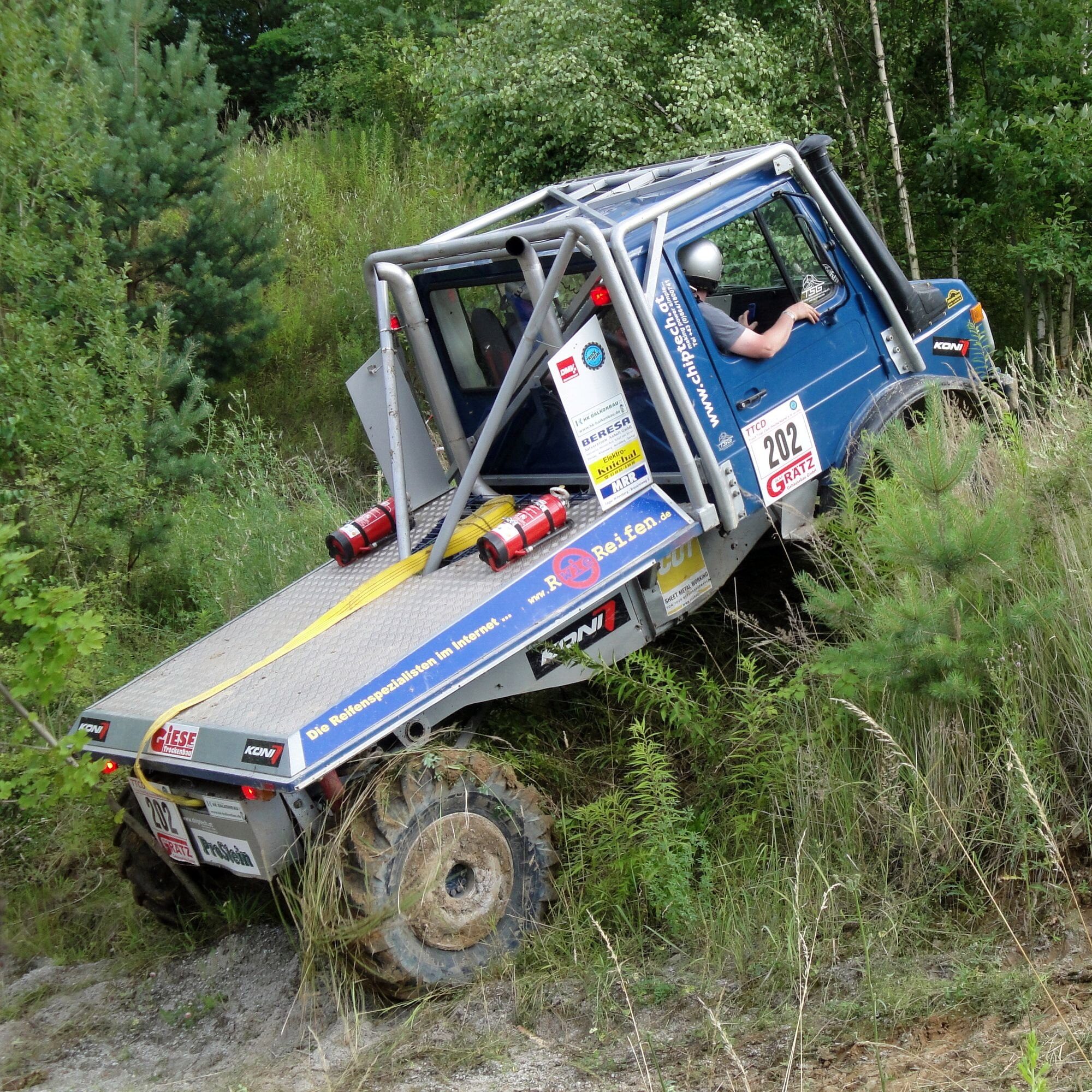 A blue, roll-caged Unimog 1300 L truck-trial competitor climbing out of a ditch on a forested course, its flatbed tilted steeply
